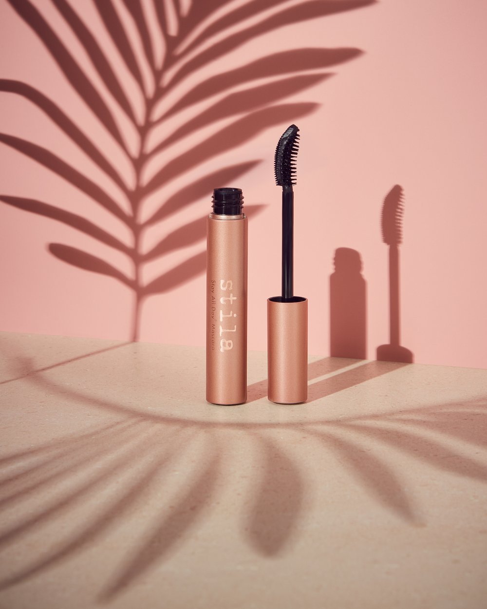 Stila mascara tube and wand on a light surface with a pink backdrop, casting a leaf shadow. Beauty product shot in a studio.