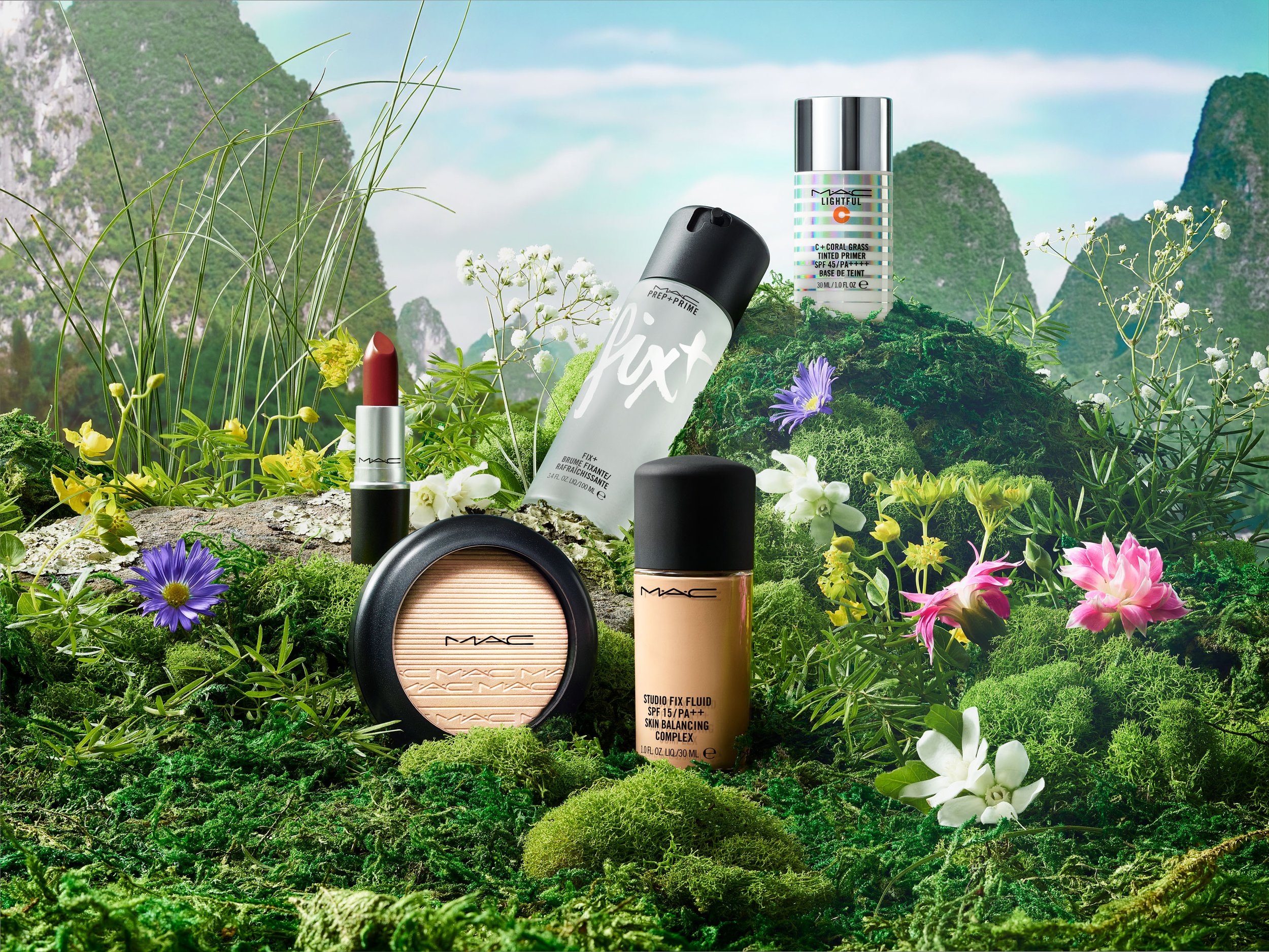 MAC makeup products, including lipstick, powder, primer, and foundation, arranged on a mossy surface with a mountain backdrop.