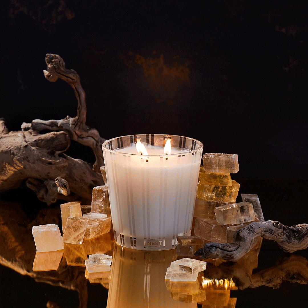 Illuminated Nest candle surrounded by decorative translucent amber and white rocks on a glossy reflective surface against a dark backdrop.