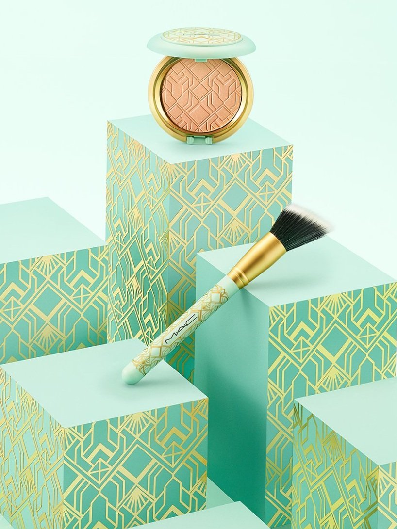 Elevated studio shot of mint and gold MAC powder compact and makeup brush arranged on patterned blocks against a mint background.