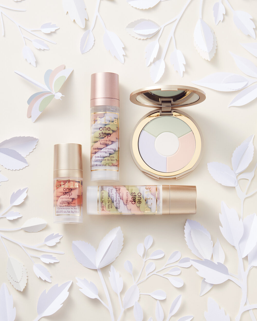 Beauty product still life of cosmetic powder compact and primer bottles with pale, soft color palette and decorative paper foliage.