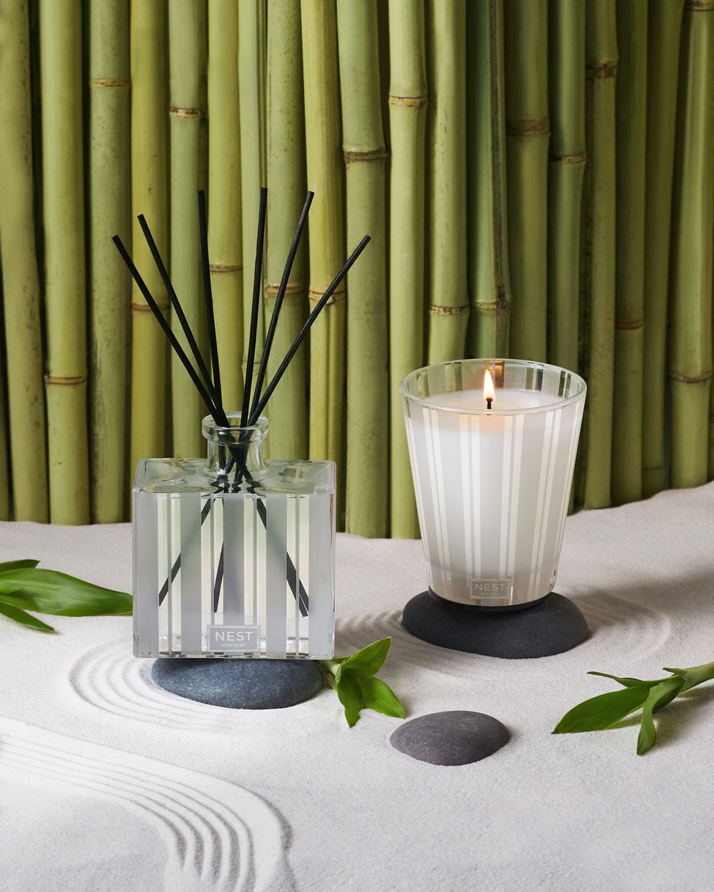 Aromatic reed diffuser and candle set on white sand with a backdrop of bamboo stalks for a relaxing zen-like ambiance.