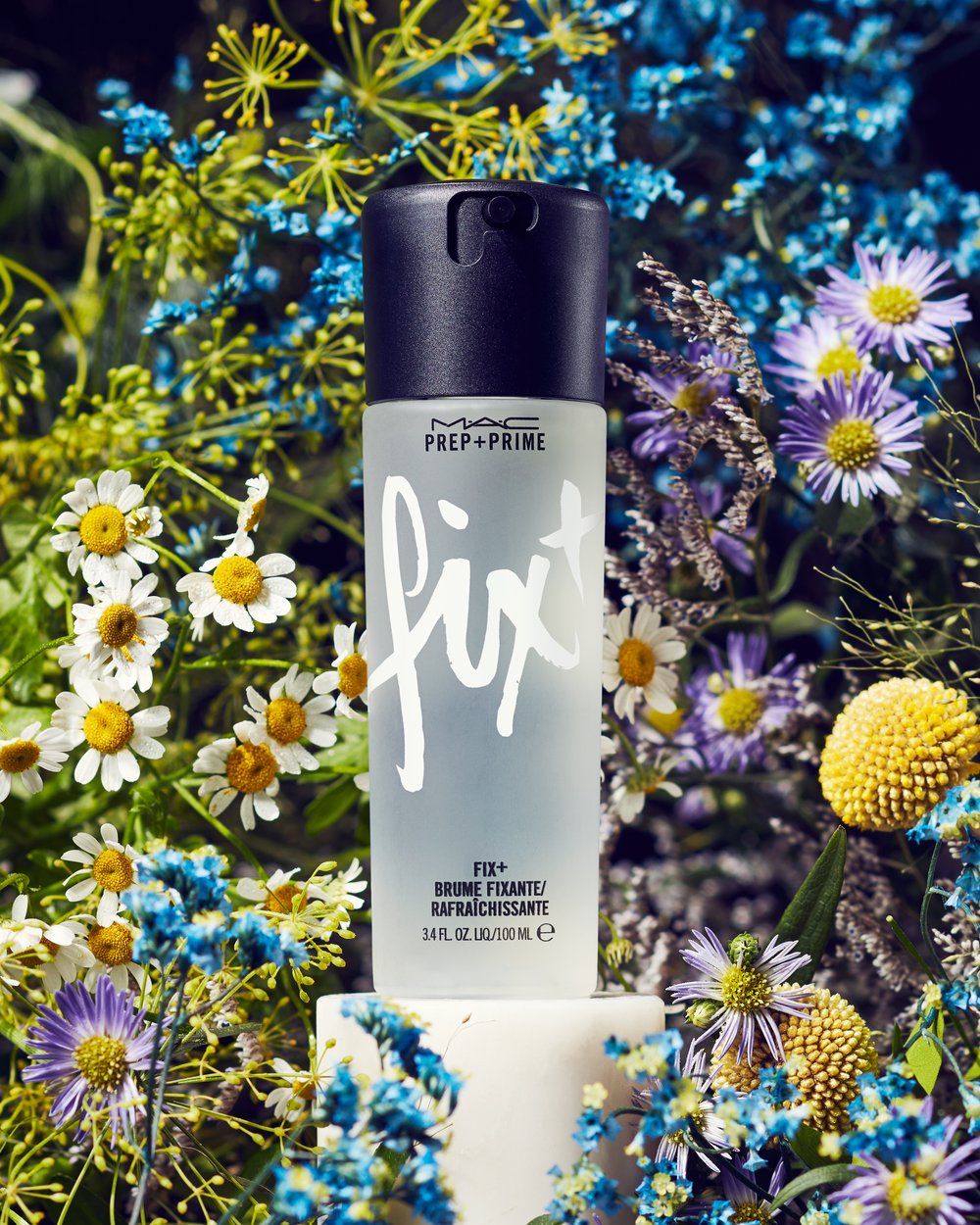 Close-up of MAC Prep+Prime Fix+ setting spray amidst daisies and other wildflowers on a white cube. Cosmetic beauty product shot.