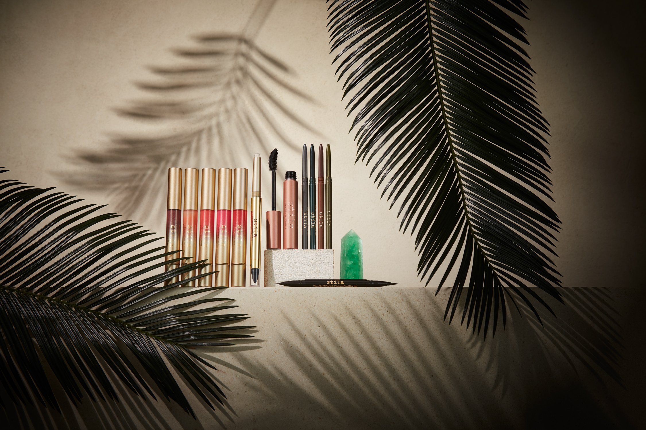 Still life of Stila brand makeup products including lip gloss, mascara, and eyeliner in gold packaging, set against a neutral background.