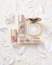 Flat lay of cosmetic bottles and a colour correcting compact surrounded by intricate paper-cut leaves.