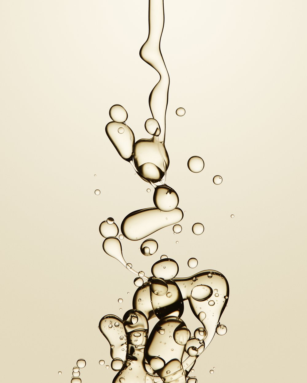 Close-up shot of translucent liquid droplets, merging and suspended against a pale beige background, creating an abstract, organic form.