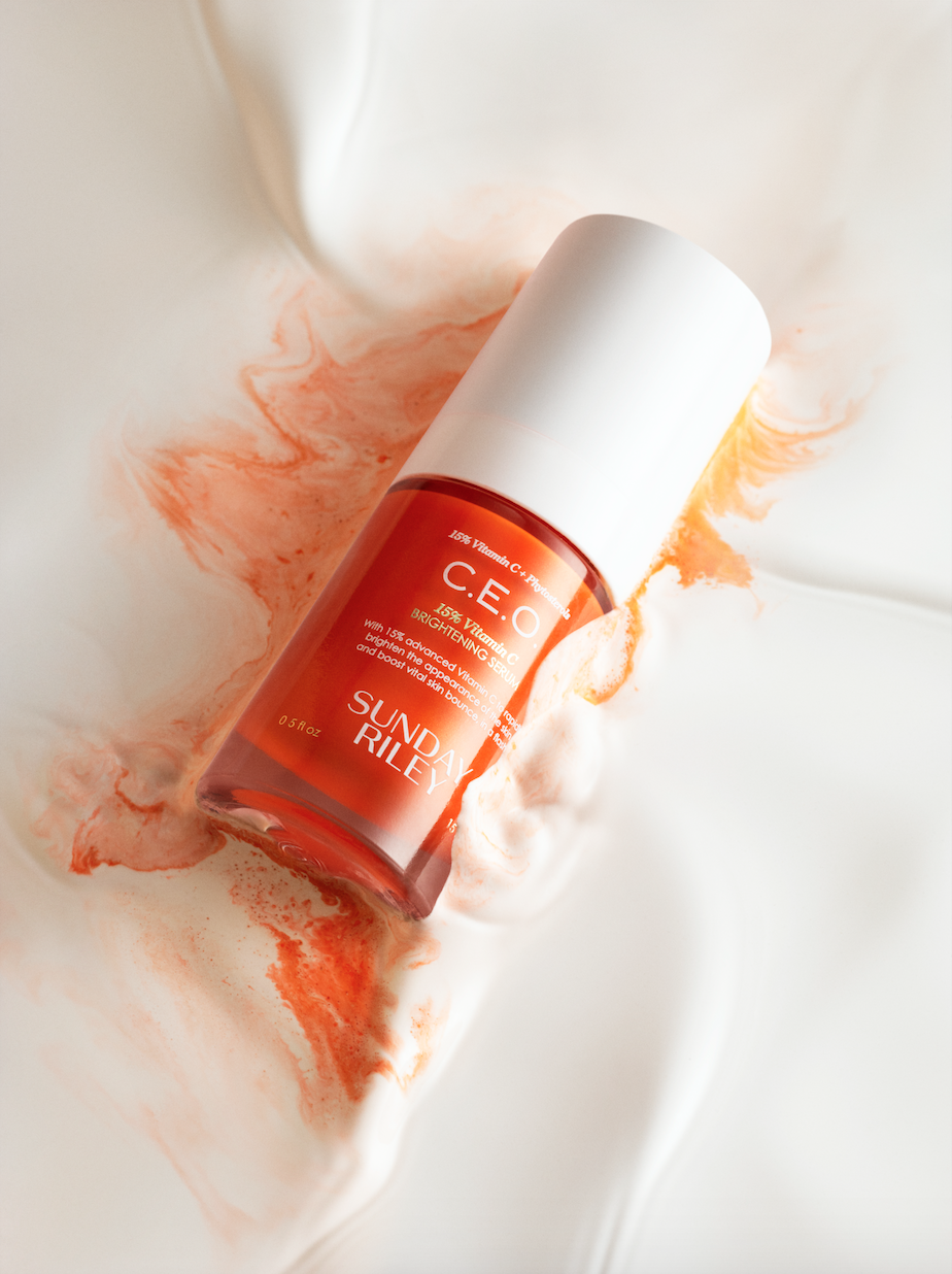 Elevated product shot of Sunday Riley C.E.O. 15% Vitamin C Brightening Serum on white satin with orange pigment. Beauty industry.