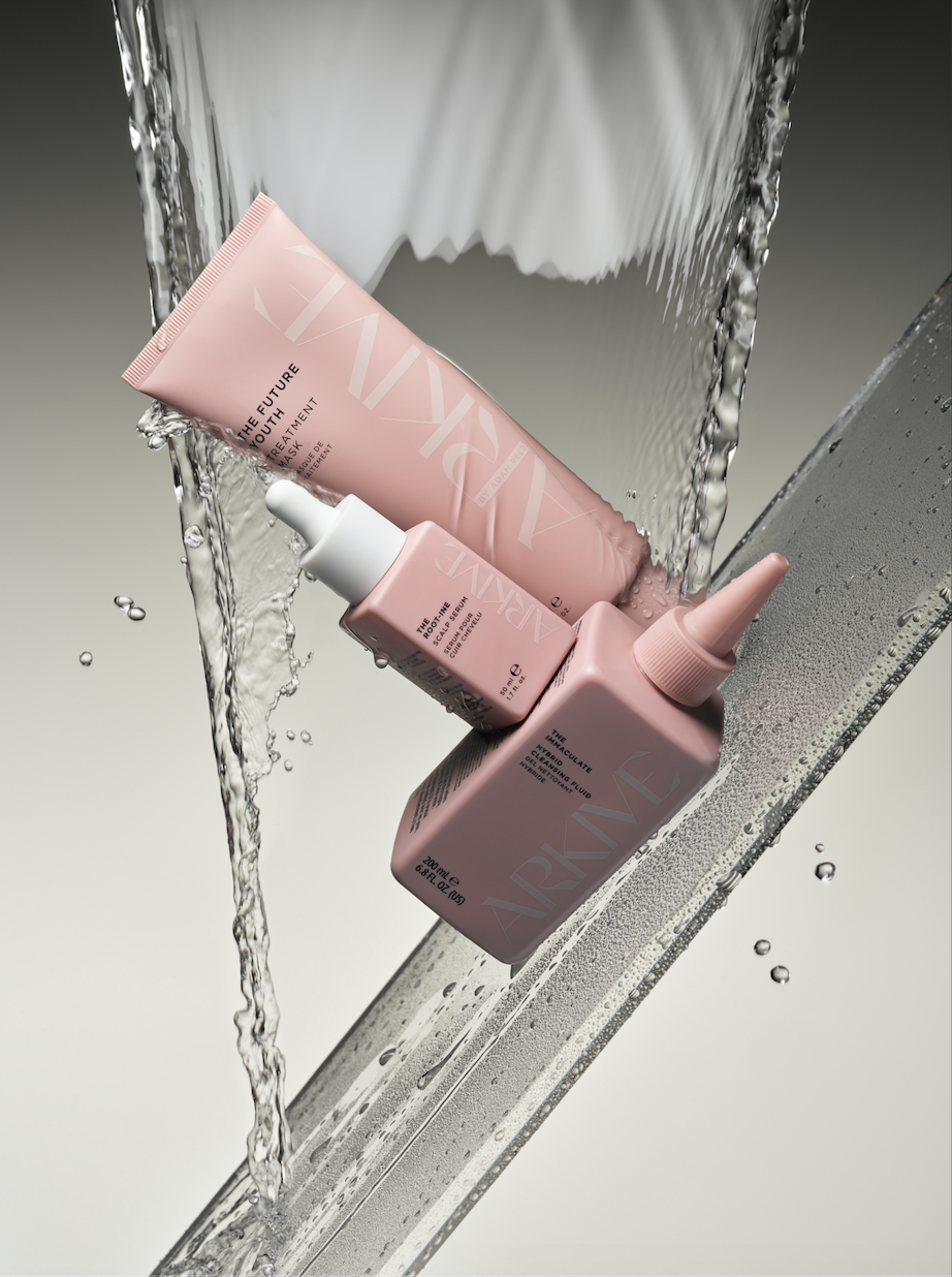 Three pink beauty product containers, a tube and two bottles, are arranged on a surface with water splashing around them.