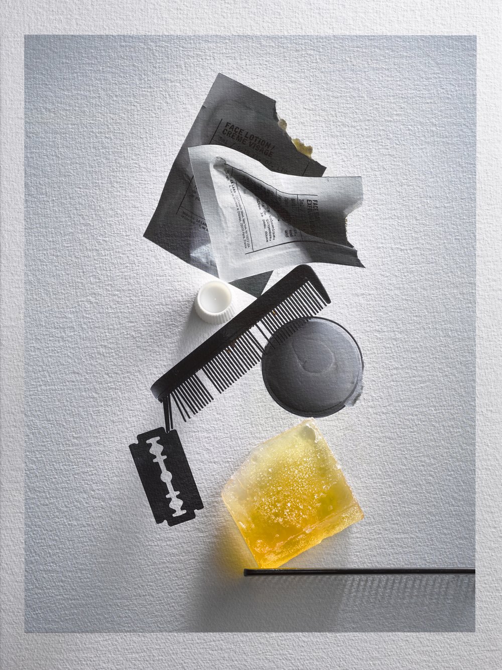 Still life shot of grooming products including a razor blade, soap, comb, cream, and face lotion on a white surface with shadows.