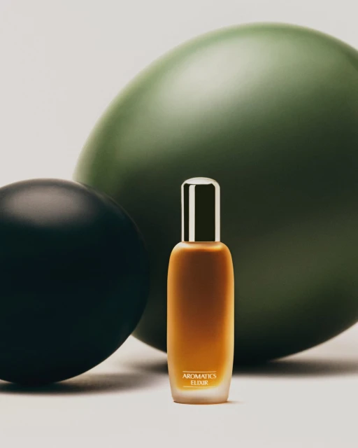 Aromatics Elixir bottle stands in front of green and black spheres on a white surface in a product shot with neutral lighting.