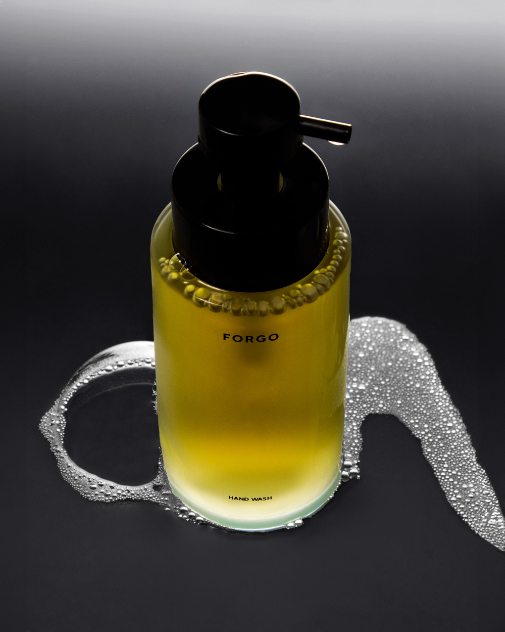 Still shot of a clear glass soap dispenser with a black pump, filled with yellow liquid, sitting on a black surface with soap bubbles.