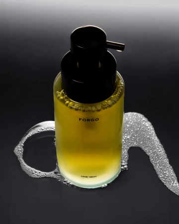 Still shot of a clear glass soap dispenser with a black pump, filled with yellow liquid, sitting on a black surface with soap bubbles.