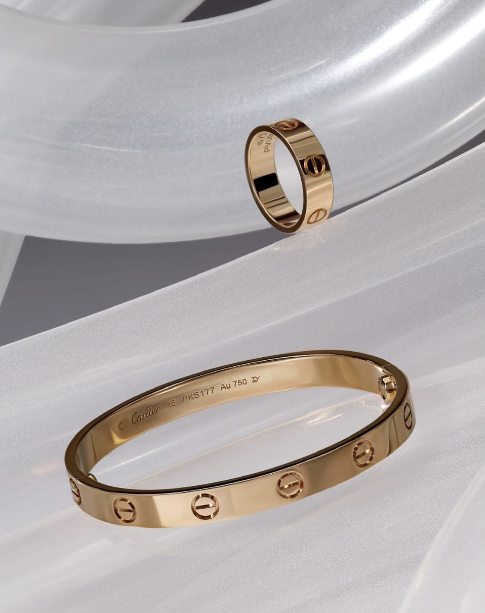 Elevated view of a gold Cartier ring and matching bangle on a white translucent surface with ambient lighting. Jewelry and accessories.