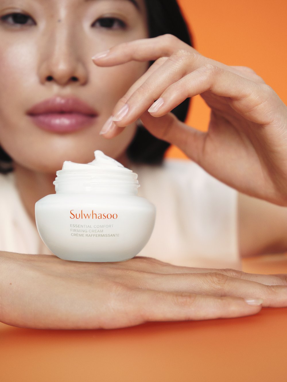 Close-up of a cream jar held by an Asian female. The jar is creamy white with orange text on a matching orange background.