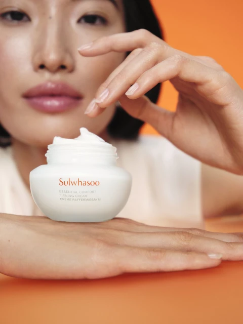 Close-up of a cream jar held by an Asian female. The jar is creamy white with orange text on a matching orange background.