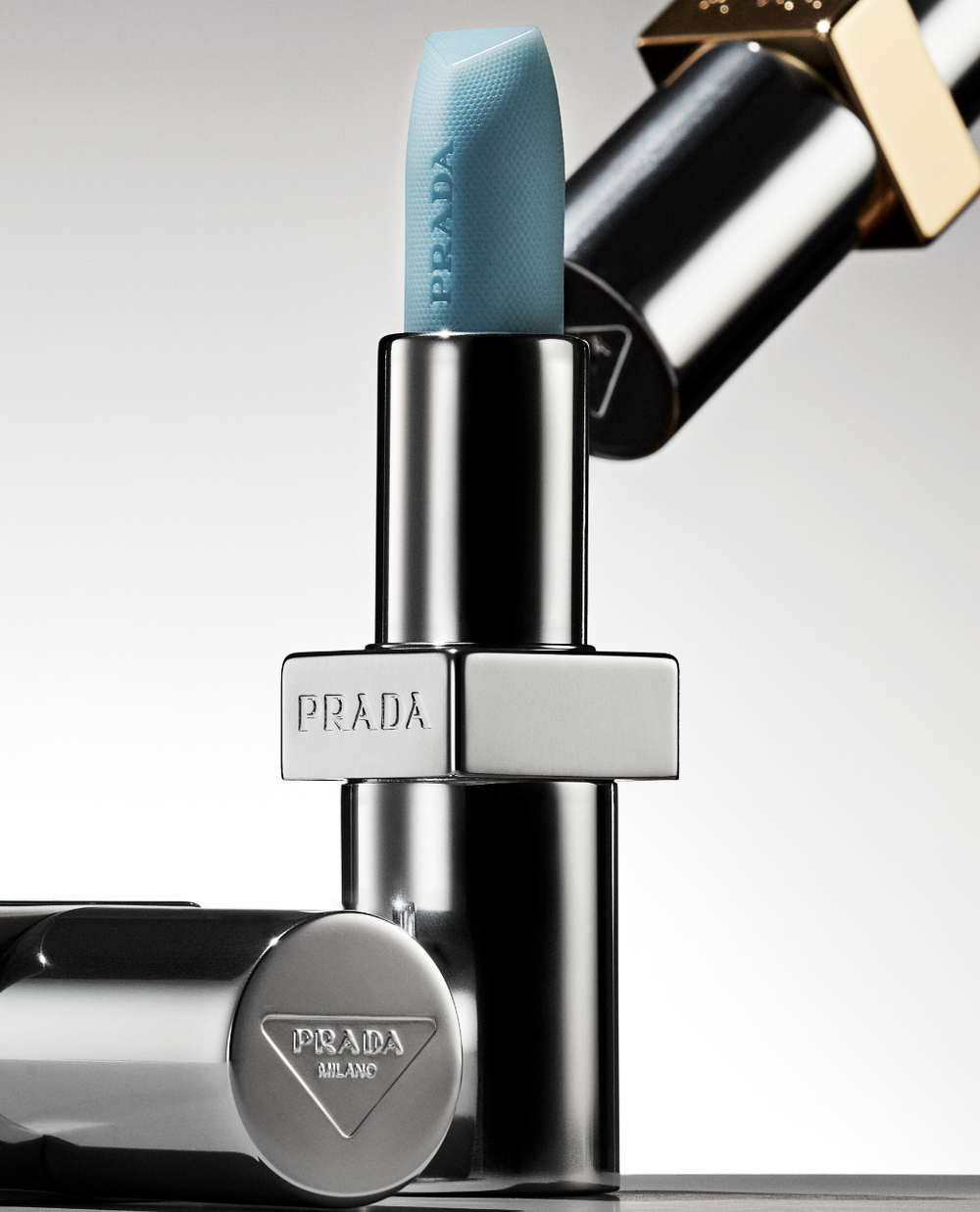 Close-up of teal Prada lipstick with silver and gold hardware against a light background, showcasing makeup and cosmetic product details.