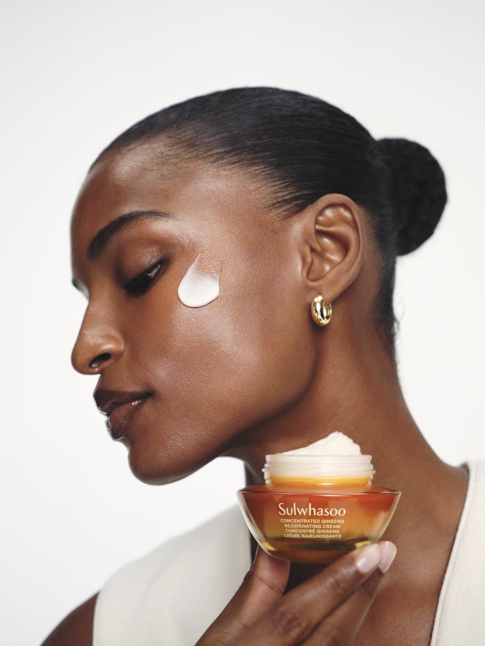 Close-up of an adult woman with skincare cream on her face, holding a jar of Sulwhasoo Concentrated Ginseng Rejuvenating Cream.
