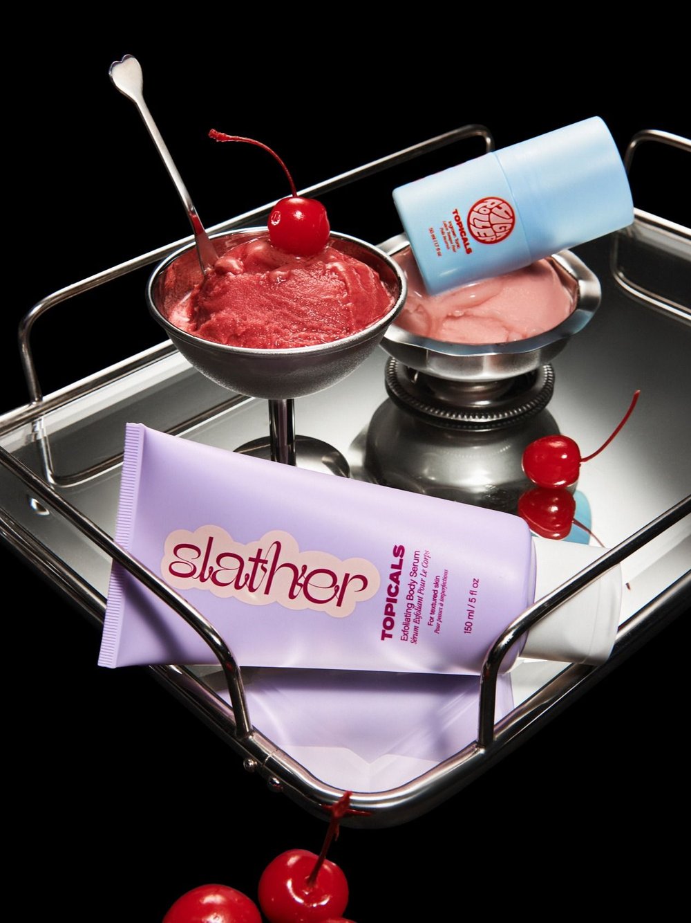 Beauty product still life on a silver tray features skincare containers, ice cream dessert in silver bowls, and bright red cherries.