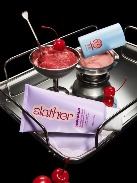 Beauty product still life on a silver tray features skincare containers, ice cream dessert in silver bowls, and bright red cherries.