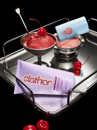Beauty product still life on a silver tray features skincare containers, ice cream dessert in silver bowls, and bright red cherries.