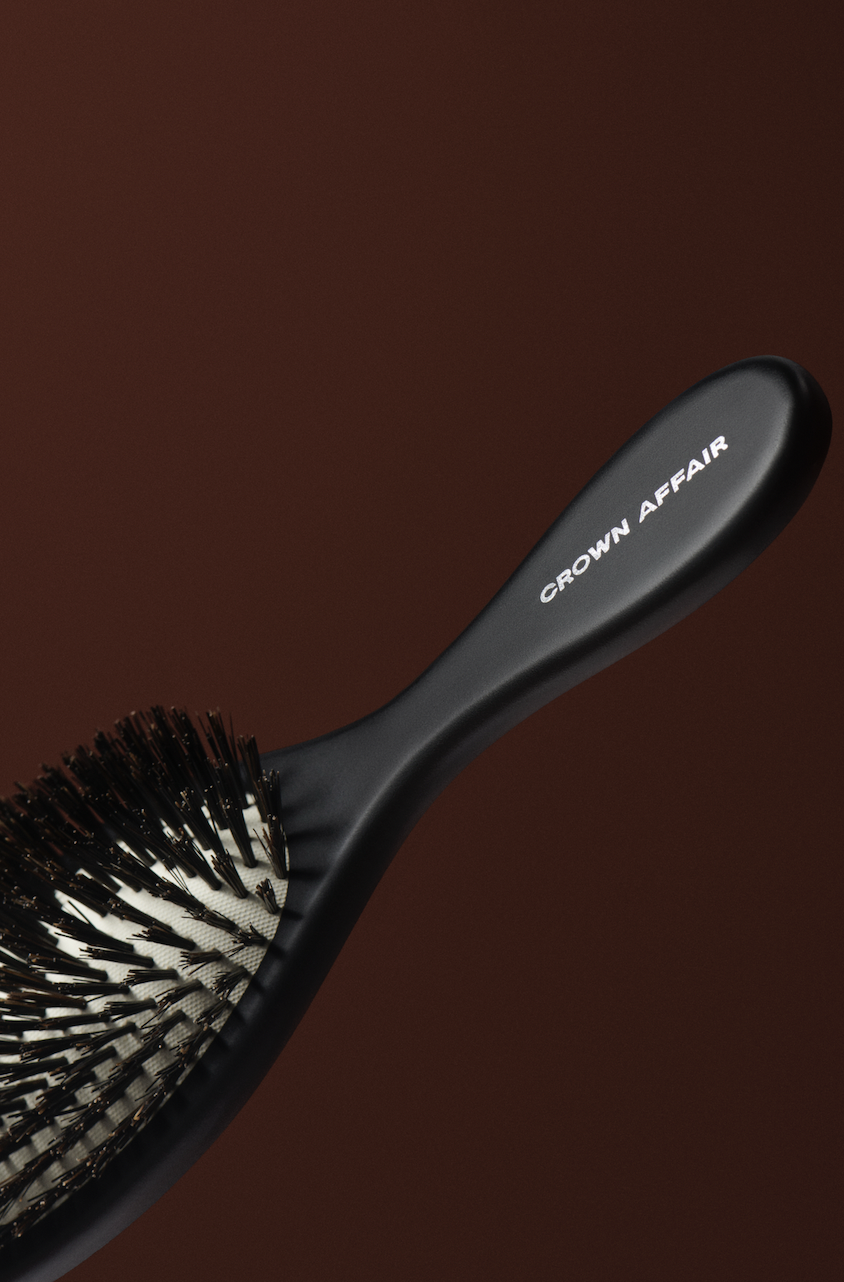 Angled close-up of a black Crown Affair hairbrush with densely packed bristles, set against a blurred brown gradient backdrop.