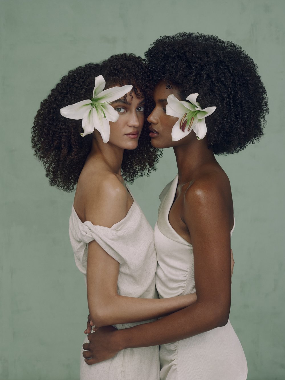 Two women with afro hairstyles and floral accents pose intimately against a muted green backdrop, wearing light-colored dresses.