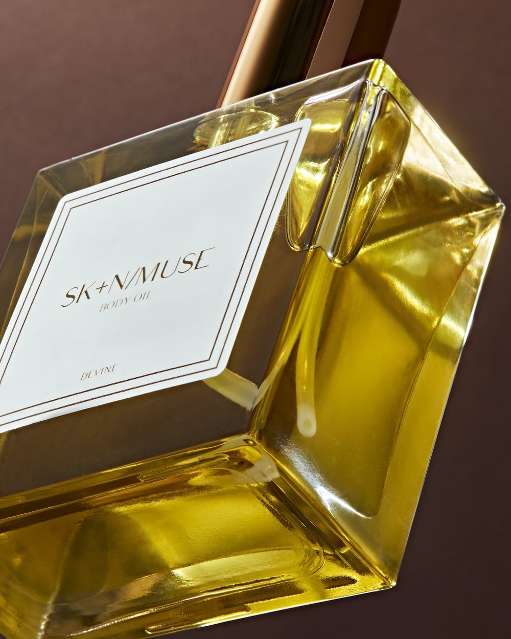 A studio shot features a clear bottle of SK+N/MUSE body oil with gold liquid, set against a dark brown background.
