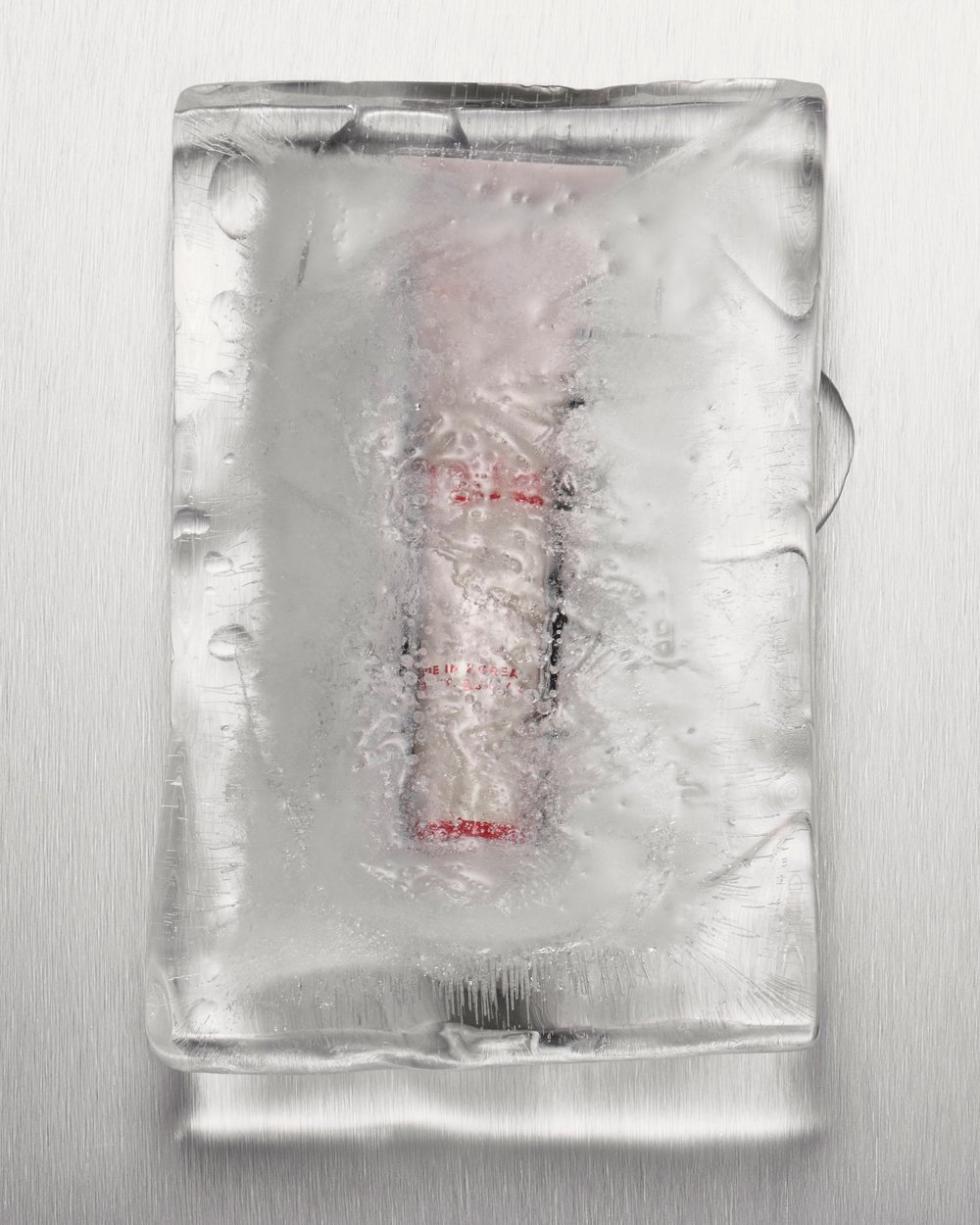 Close-up of an ice block encasing an unidentified object, set against a brushed metal background with condensation and textural ice details.