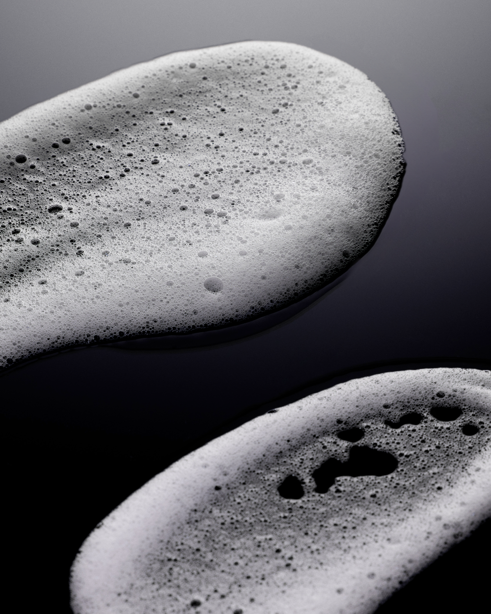 Close-up studio shot of white soap foam on a dark, reflective surface. Abstract beauty and cleanliness concept.