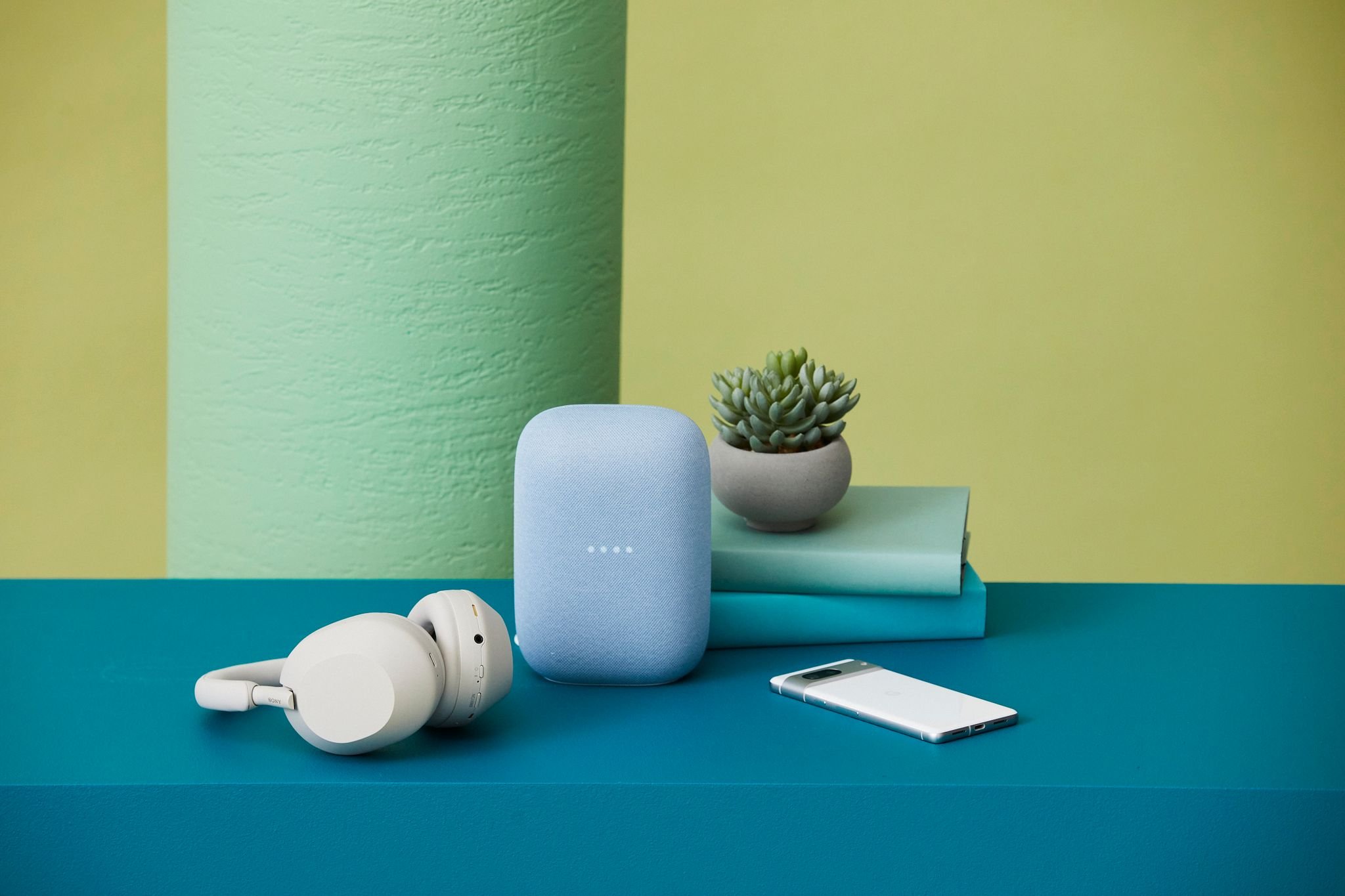 Still life featuring a blue Google Nest speaker, white headphones, grey succulent, books, and white cell phone against green background.