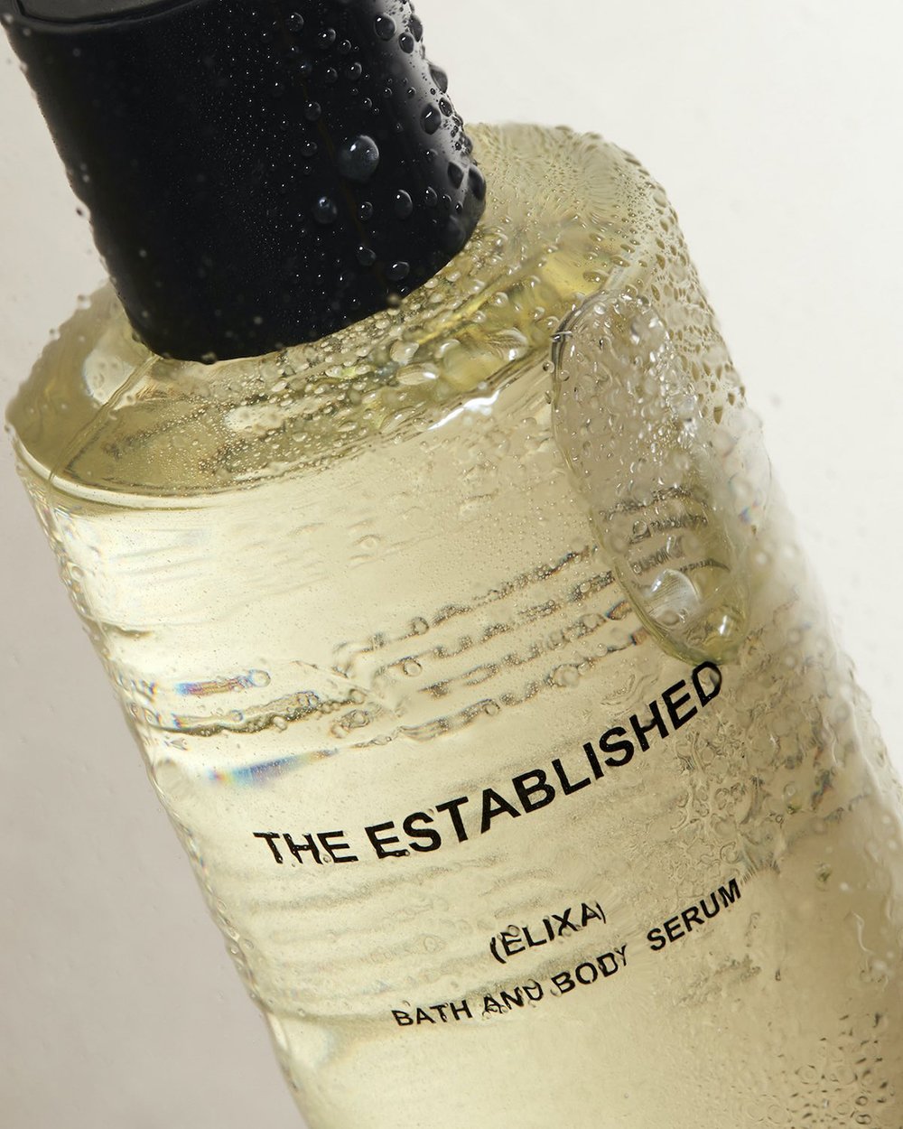 Close-up of a bottle of clear bath and body serum with a black cap, featuring visible water droplets and minimal white background.
