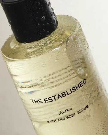 Close-up of a bottle of clear bath and body serum with a black cap, featuring visible water droplets and minimal white background.