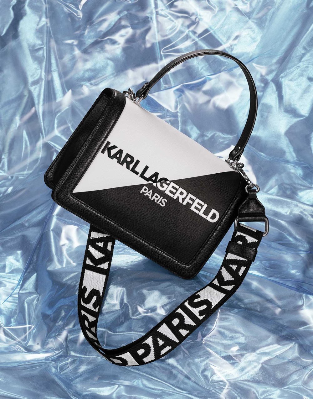 Karl Lagerfeld black and white purse featuring a strap with the brand's name, set against a textured, iridescent blue background.
