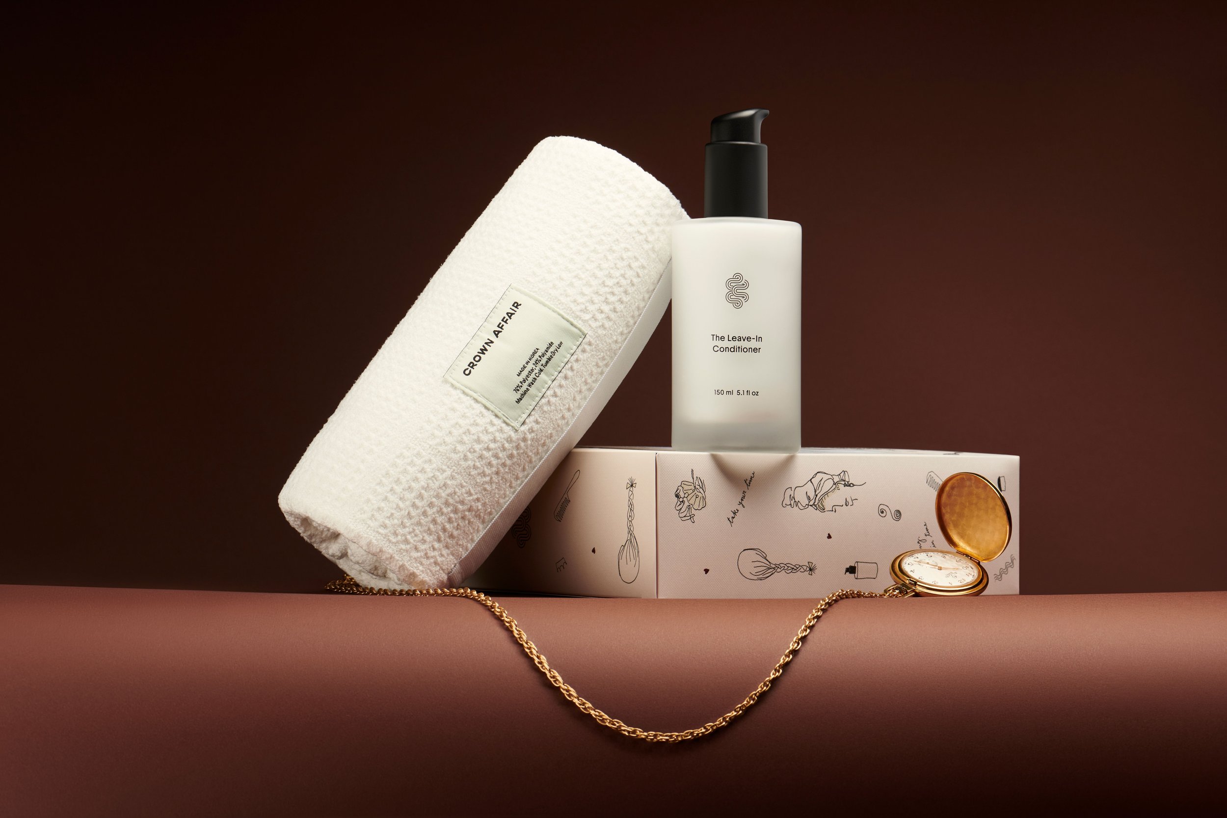 Hair product bottle, white towel, and gold necklace on brown background in an indoor studio shot. Beauty product still life.