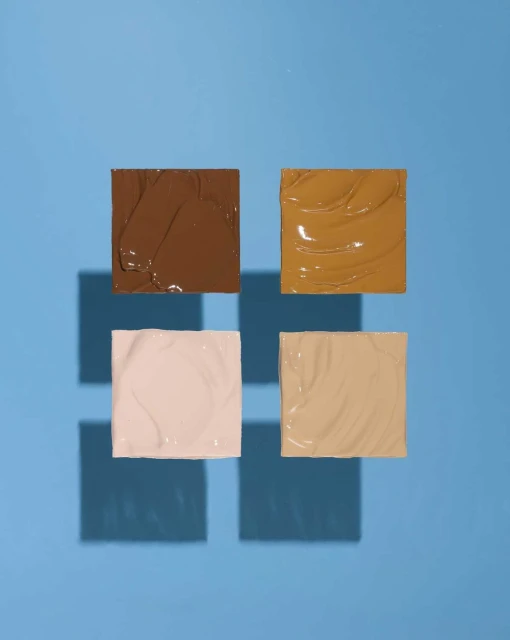 Four squares of beige and brown cosmetic cream foundation shades on a blue background with square shadows in a simple product shot.