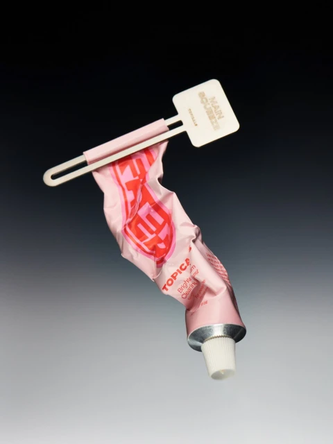 Close-up of a pink tube of topical cream held by a white plastic squeezer against a dark grey gradient background in a studio shot.