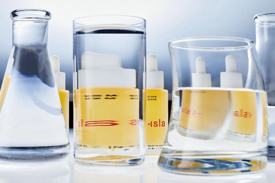 Product shoot featuring Isla skincare: yellow cream and serum bottles partially submerged in glass beakers filled with water in studio setting.
