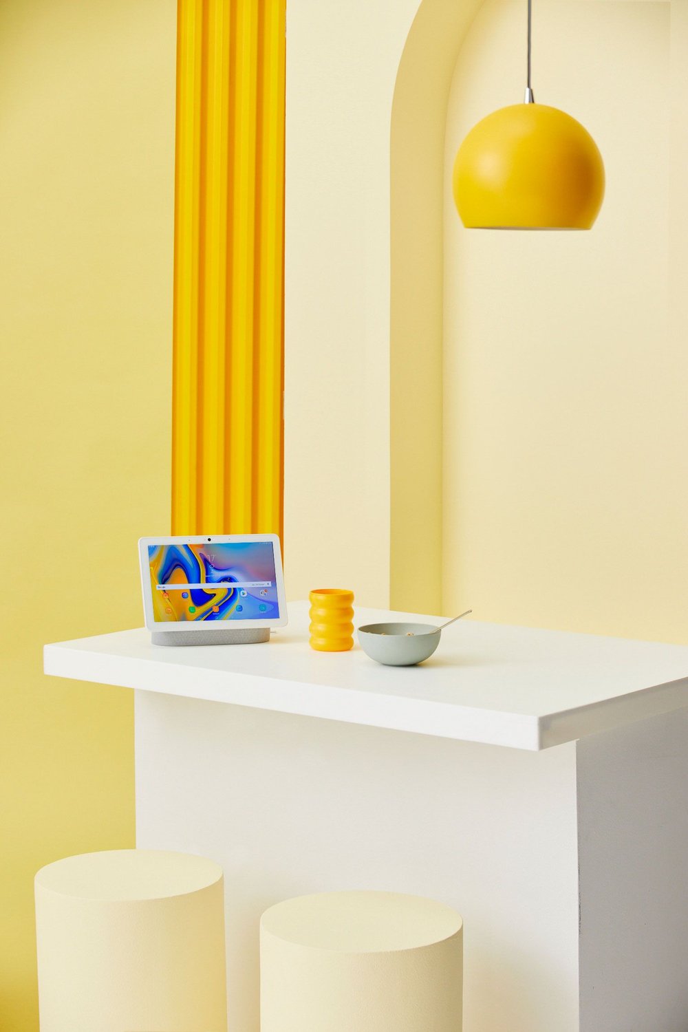 Minimalist studio shot features a laptop on a white counter, complemented by a bowl, decorative yellow objects, and a hanging pendant lamp.