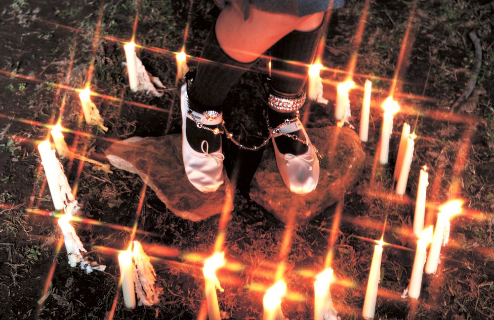 Ballet slippers on a rock surrounded by lit candles outdoors creating a dramatic, warm-toned, soft-focus, and mystical scene.