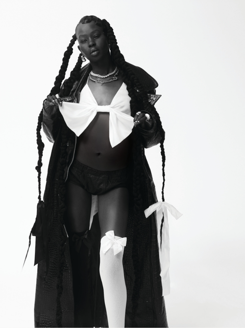 A striking studio shot of a Black adult female model wearing a black coat with ribbons and long braids, set against a clean, white backdrop.