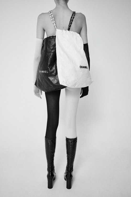 Back view of a slender figure showcasing a two-tone bag with a 'CHANEL' logo, tights, high-heeled boots, and opera gloves on a plain background.