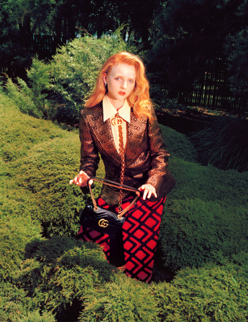 Red-haired woman in a patterned blazer and pants poses in lush greenery, holding a black velvet Gucci purse with a gold chain.