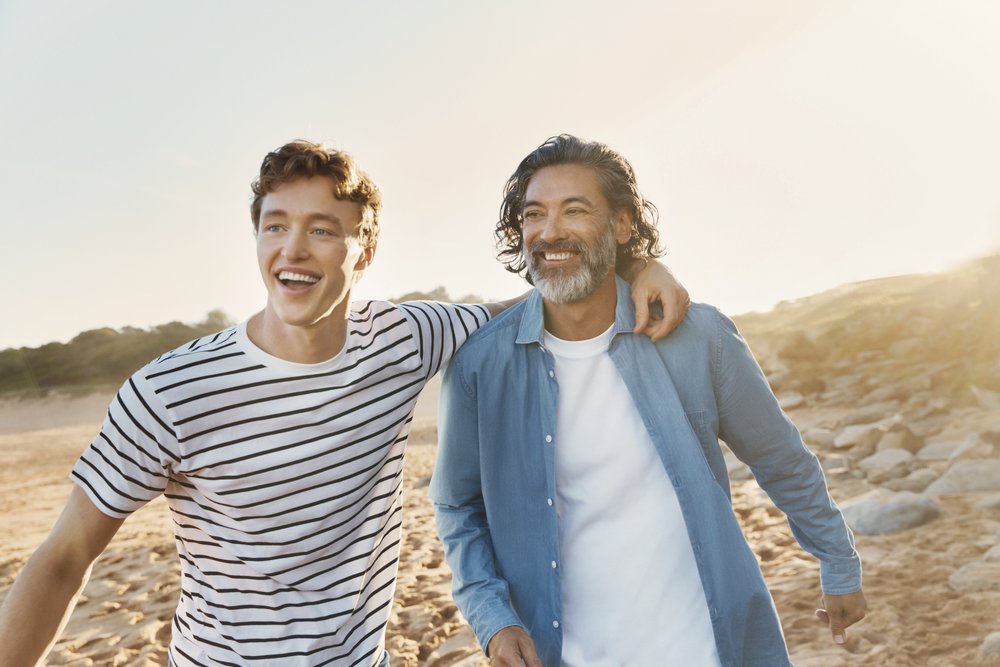 Two men, a father and son, smile while walking arm-in-arm on a sunlit beach. The younger man wears a striped shirt and the older man a button-down.