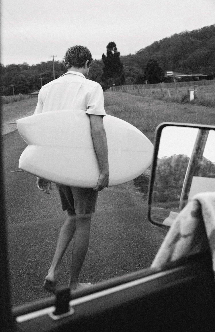 Black and white shot of a man walking away from the camera carrying a surfboard, outdoors on a road, trees and hillside in background.