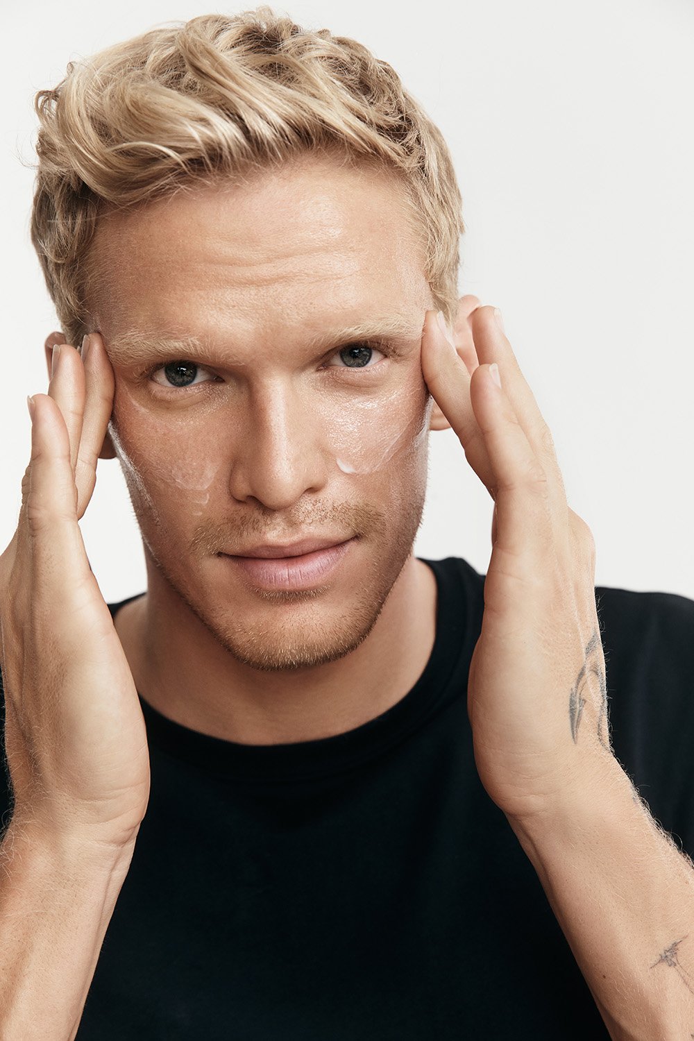 Close-up of a blonde male applying facial cream to his face, set against a neutral background, with a focus on skincare and grooming.