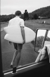 Man carrying a surfboard walking along a rural road, viewed from inside a car by Ming Nomchong