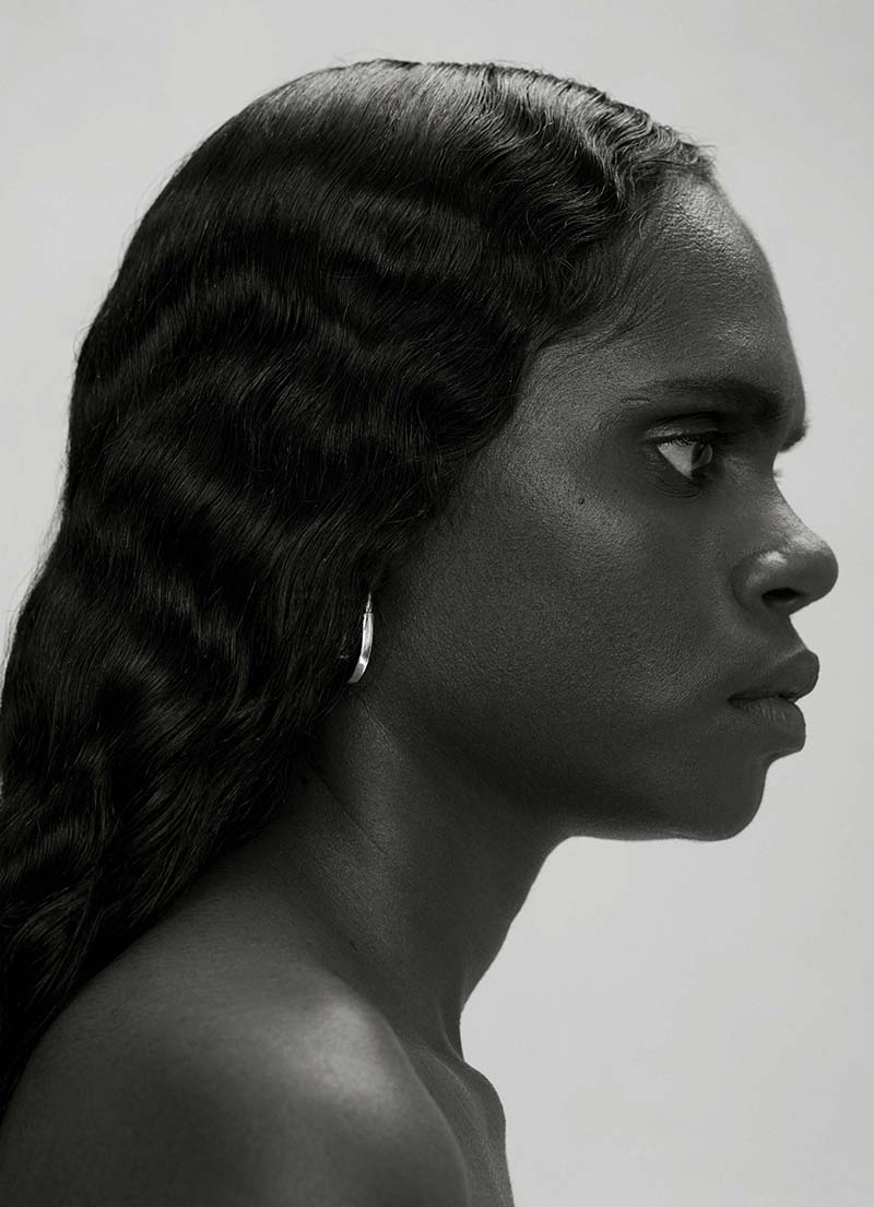 Monochrome portrait of a dark-skinned woman in profile, showcasing wavy hair, subtle jewelry, and a stark, contrasting background.