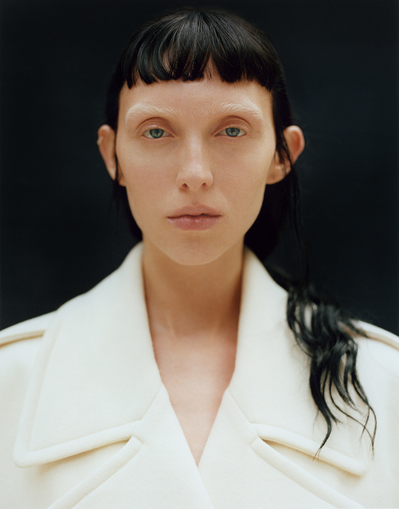 Close-up studio portrait of a woman with dark hair and a white coat against a dark background, showcasing a minimalist fashion aesthetic.