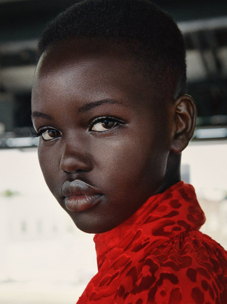 Close-up portrait of an African woman with short dark hair, wearing a red patterned top, against a blurred neutral background.