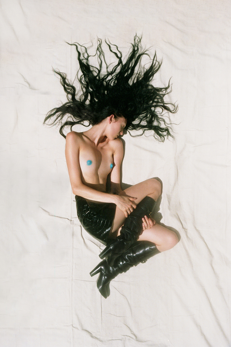 A woman with long, dark hair and black boots lies on her back on a white fabric surface, photographed in bright studio lighting.